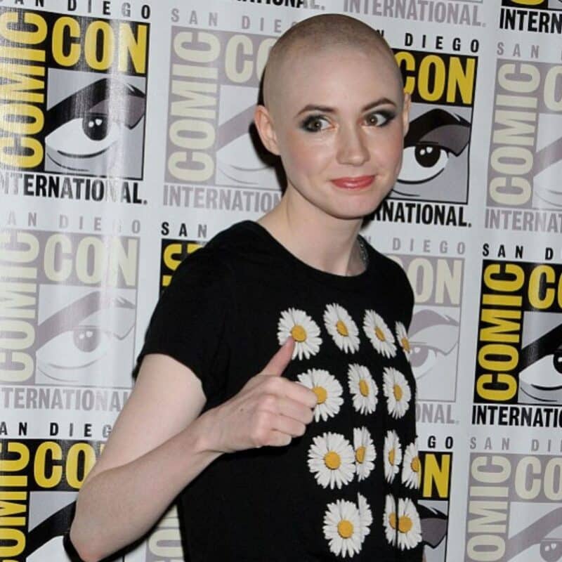 Karen Gillan, bald, giving a thumbs-up at San Diego Comic-Con, wearing a black shirt with daisy prints