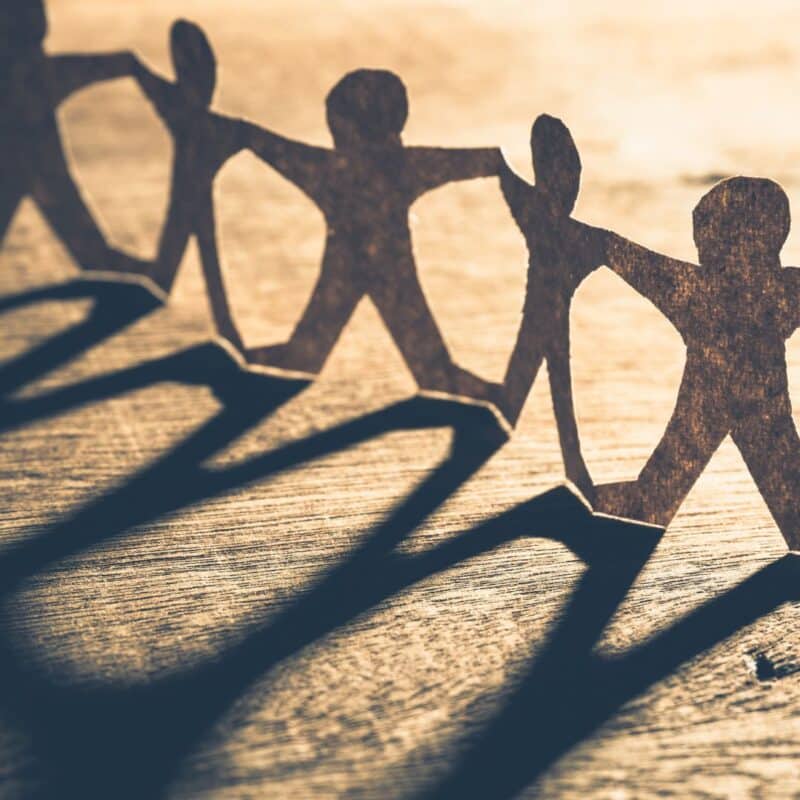 Paper cutouts of human figures holding hands, casting long shadows on a wooden surface