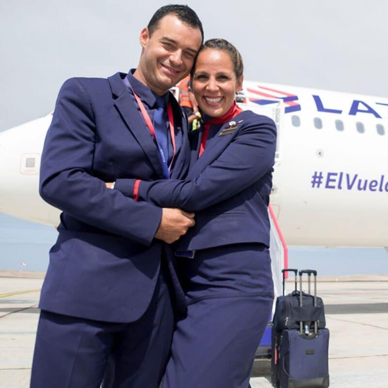 Two smiling flight attendants in uniform embrace in front of a LATAM airplane
