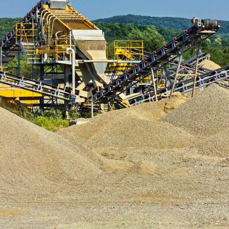 Large piles of industrial sand and gravel near a processing facility with conveyor belts and steel structures, set against a backdrop of trees and hills