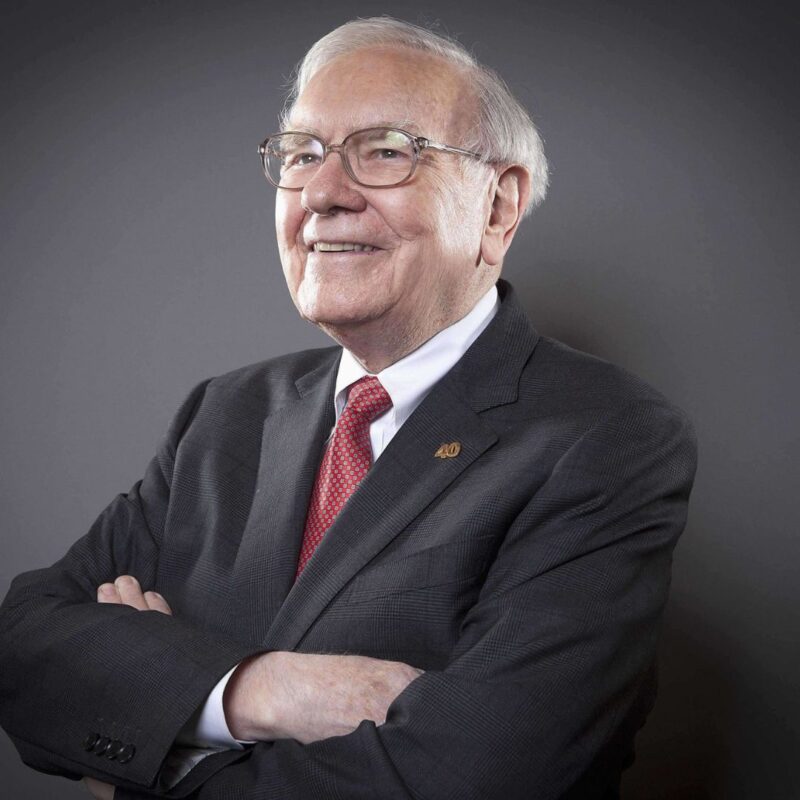 Warren Buffett wearing a dark suit with a red tie and smiling with his arms crossed