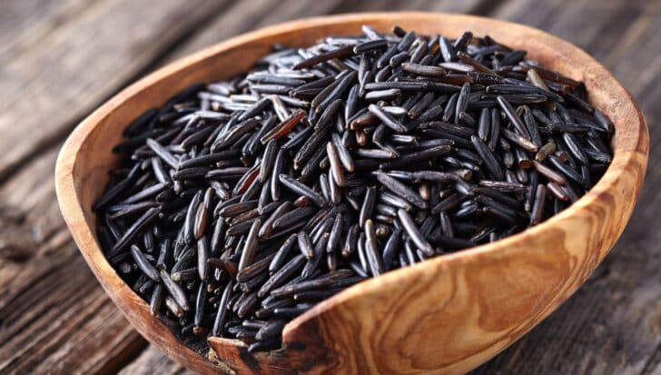 A wooden bowl filled with wild rice grains on a wooden surface