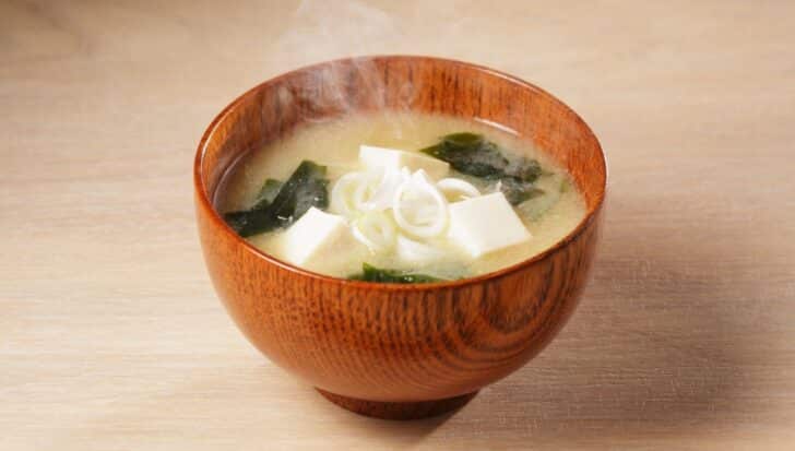 A wooden bowl filled with steaming miso soup containing tofu cubes and sliced green onions