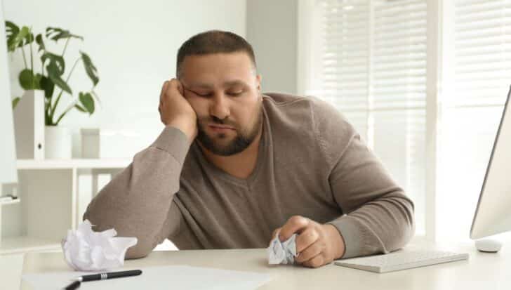 A person looking bored and disengaged at a desk, surrounded by a computer, crumpled paper, and a pen.