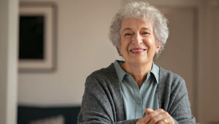 Smiling older woman with curly gray hair wearing a gray sweater over a light blue shirt