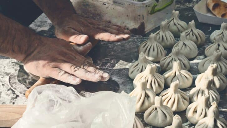 Hands shaping dough on a floured surface with multiple uncooked Georgia Khinkali nearby