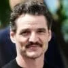 Pedro Pascal with short, wavy hair and a mustache smiling outdoors with blurred greenery in the background