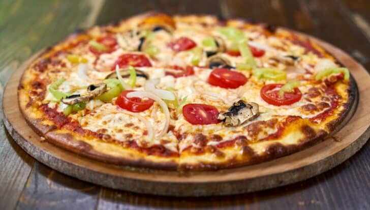 A pizza with toppings like tomatoes, mushrooms, onions, and green peppers on a wooden serving board