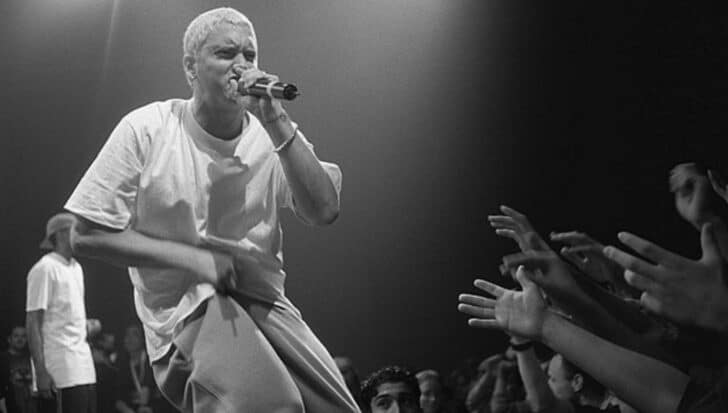 Eminem with a microphone is on stage in front of a crowd with hands reaching out
