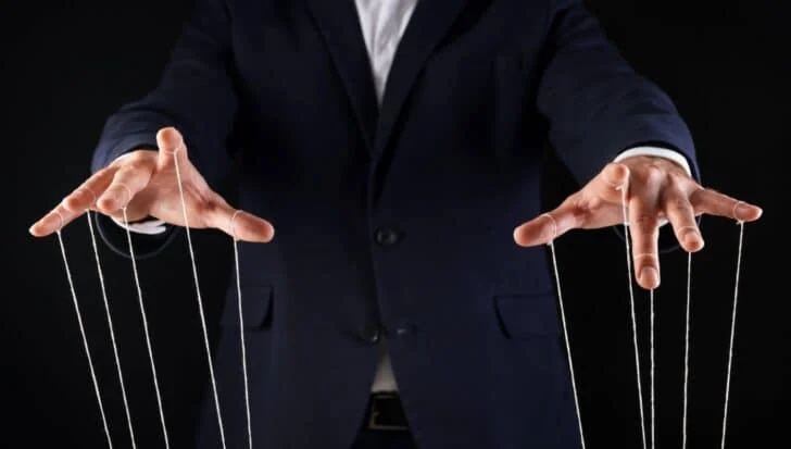 A person in a suit manipulates strings like a puppeteer, symbolizing control and possibly secretive experiments