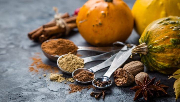 Various spices including ground cinnamon, cloves, nutmeg, and star anise with small pumpkins in the background