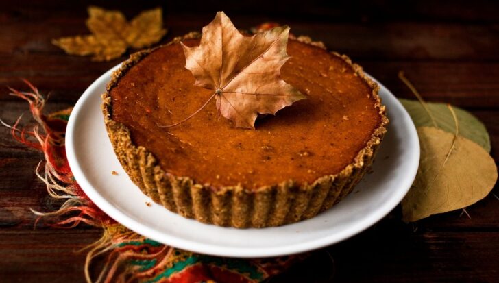 Pumpkin pie on a white plate, topped with a dry maple leaf, on a wooden table with autumn leaves and a colorful fabric nearby