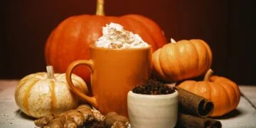 A warm setting with an orange mug topped with whipped cream, surrounded by pumpkins, ginger, cinnamon sticks, cloves, and a small bowl of spices